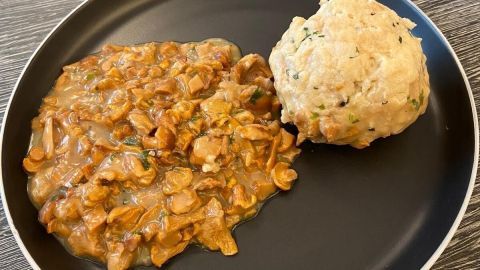 Eine cremige Schwammerlsoße liegt auf einem dunklen Teller, neben einem knusprigen Knödel in einem modernen Essumfeld.