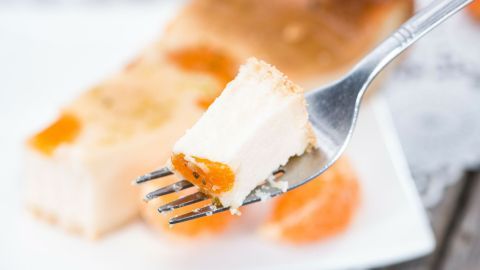 Ein Stück Mikrowellen-Käsekuchen liegt auf einem weißen Teller, während eine Gabel einen Bissen mit Aprikosenstücken aufnimmt.