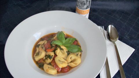 Eine Schüssel mit Tortellini in Tomatensauce, garniert mit frischem Spinat und Basilikum, liegt auf einem blauen Tisch.