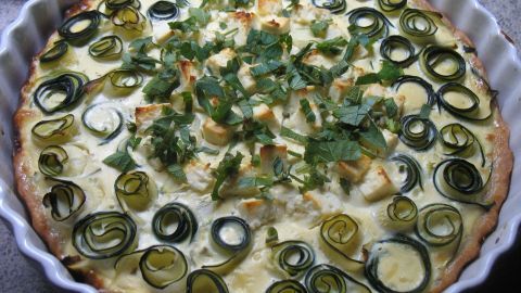 Eine frisch gebackene Zucchini-Tarte mit Käse und Kräutern liegt in einer runden Form, dekoriert mit grünen Zucchinischeiben.