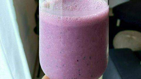 Ein rosa Buttermilch-Drink mit gefrorenen Heidelbeeren wird in einem Glas gehalten, während man in einem modernen Raum steht.