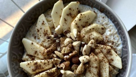Ein Schüssel Apfel-Nuss Porridge mit Bananenscheiben und Nüssen, auf einem Tisch neben einem Fenster.