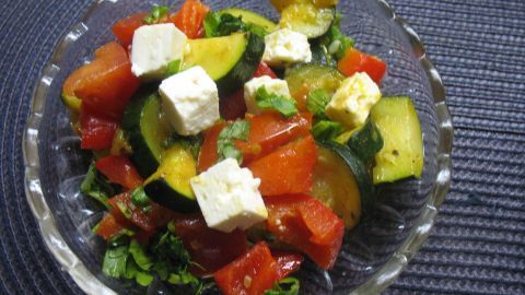 Ein bunter Gemüse-Kräuter-Salat mit fetakäse liegt in einer Glasschüssel und strahlt Frische auf einem blauen Untergrund aus.
