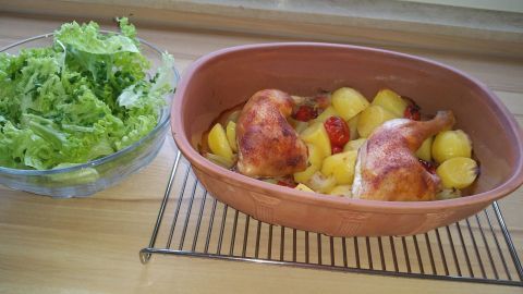 Zarte Hähnchenschenkel garen mit goldbraunen Kartoffeln und Cocktailtomaten im Römertopf, umgeben von frischem Salat.