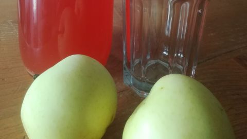 Ein Glas und eine Flasche Apfelsirup stehen auf einem Tisch, umgeben von frischen Äpfeln in einem gemütlichen Raum.