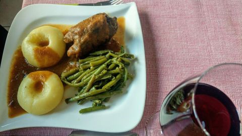 Schlesische Kartoffelklöße liegen neben einem saftigen Fleischstück und grünen Bohnen auf einem weißen Teller. Ein Glas Wein ergänzt das Mahl.