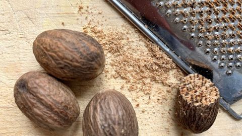 Muskatnüsse liegen auf einem Holzbrett, während eine Reibe daneben feines Muskatnuss-Pulver zeigt, das frisch gerieben wird.
