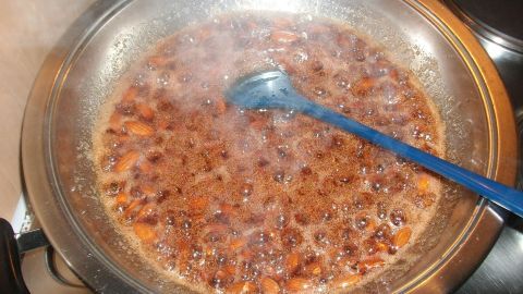 In einer Pfanne kochen gebrannte Mandeln in einer süßen, dampfenden Zuckermischung, während ein blauer Löffel umrührt.