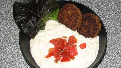 Dunkle Frikadellen liegen neben einer cremigen Buchweizen-Creme, garniert mit frischen Tomatenstücken, auf einem schwarzen Teller.