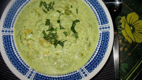 Eine grüne Sommersuppe befindet sich in einer dekorativen Schüssel, garniert mit frischen Kräutern, auf einem dunklen Tisch.