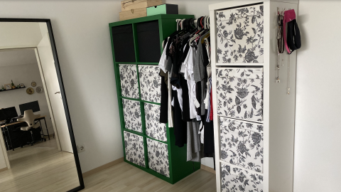 Zwei Kallax Regale stehen in einem modernen Raum, eines grün und eines weiß mit blumigen Motiven, beide organisieren Kleidung und Accessoires.