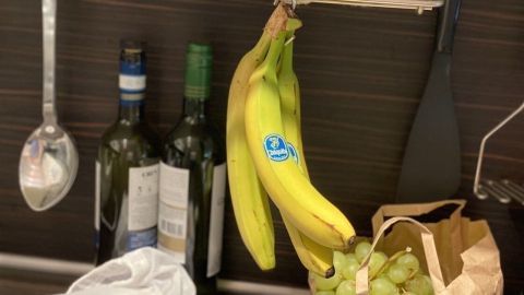 Gelbe Bananen hängen in einer Küche über einem Regal, während Trauben in einer Papiertüte neben einer Flasche stehen.