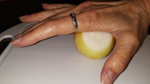 Eine Hand hält eine geschälte Zwiebel auf einem Schneidebrett, während eine Klinge zum Schneiden bereit ist.