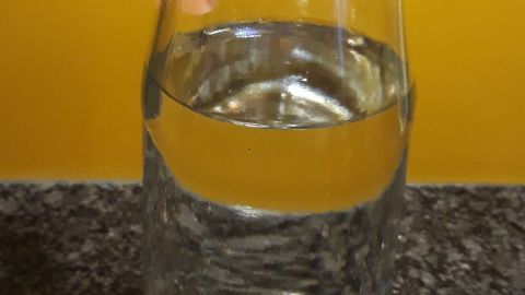 Ein Glas mit Wasser steht auf einer dunklen Oberfläche, während es von einem hellen, gelben Hintergrund umgeben ist.