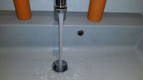 Ein Wasserhahn lässt Wasser in ein weißes Waschbecken fließen, während orangefarbene Behälter im Hintergrund stehen.