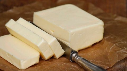 Ein Stück Butter liegt auf einem Stück Pergamentpapier, während ein Buttermesser neben den geschnittenen Portionen steht. Im Hintergrund sieht man eine braune Kaffeekanne.