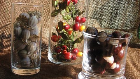 Glasbehälter mit dekorativen herbstlichen Elementen wie Beeren und Samen stehen auf einem braunen Stoffhintergrund.