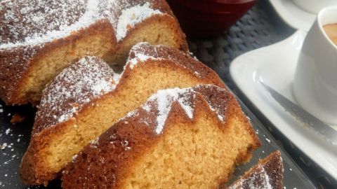 Saftiger Orangenkuchen, in Stücke geschnitten, auf einem schwarzen Teller neben einer roten Pflanze und zwei Tassen Kaffee.