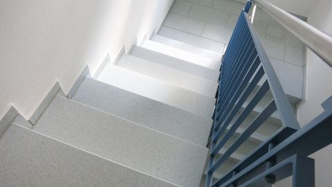 Beim Streichen von Kellertreppen diese mithilfe von Sand rutschfest machen. Kellertreppe mit einer rutschfesten Oberfläche und blauen Handlauf, die in einem hellen, modernen Raum führt.