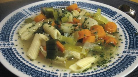 Eine bunte Karotten-Lauch-Suppe mit Gemüse und Pasta in einer dekorativen Schüssel, serviert auf einem dunklen Untergrund.