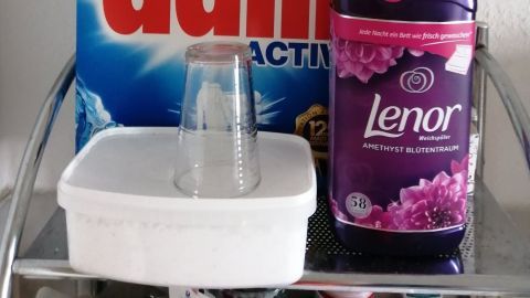Auf einem Regal stehen ein Waschpulverkarton und eine Flasche Weichspüler, die zur Wäschepflege genutzt werden.