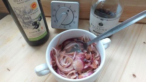 Eine Schüssel mit Raspelzwiebeln wird mit einem Löffel umgerührt, während Olivenöl und Essig als Zutaten daneben stehen.