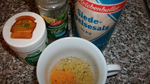 Ein Becher mit einem rohen Ei, gewürzt mit Pfeffer und Salz, steht auf einer Küchenarbeitsplatte neben Gewürzen.