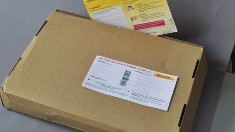Ein brauner Versandkarton liegt auf einem Tisch, beschriftet mit einem DHL-Etikett für die Paketzustellung.