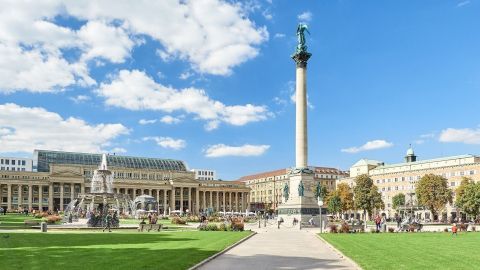 Der Platz zeigt eine monumentale Säule mit Statue, umgeben von einem lebhaften Park und historischer Architektur in Stuttgart.