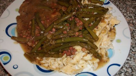 Ein Teller mit Spätzle, Bohnengemüse und Schweinehals wird appetitlich angerichtet und steht auf einer grauen Arbeitsplatte.