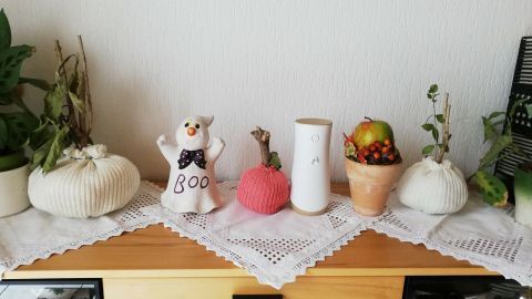Kürbisse aus Strickpullovern schmücken eine Anrichte, umgeben von Pflanzen und einer kleinen Dekofigur mit dem Text "Boo".