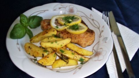 Ein Teller zeigt ein Zitronenschnitzel, garniert mit Zitronenscheiben, umgeben von gebackenen Kartoffeln und einem Basilikumblatt.