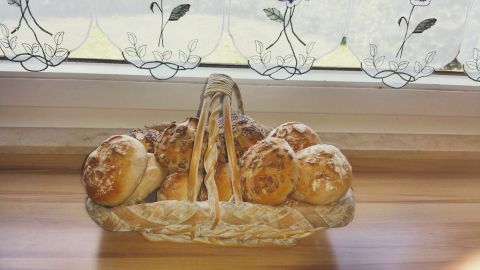 Ein Korb mit frisch gebackenen Brötchen steht auf einem Tisch unter einem Fenster mit floralem Vorhang.