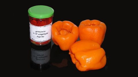 Drei orangefarbene Paprika liegen neben einem Glas mit gehäuteten Paprika in Öl, auf einem schwarzen Hintergrund.