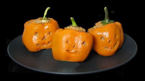 Orangefarbene, gruselig dekorierte Paprika mit Gesichtsaus schnitt liegen auf einem schwarzen Teller, gefüllt mit veganem Inhalt.
