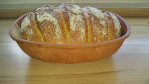 Ein frisch gebackenes Weißbrot liegt in einem römischen Tontopf, auf einer hellen Holzoberfläche, gekrönt von einer feinen Mehlschicht.
