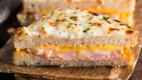 Ein goldbrauner Croque-Monsieur liegt auf braunem Papier und Holz, umgeben von Besteck und einer Essiggurke.