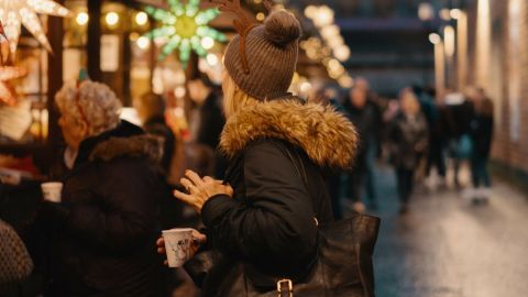 Eine Person mit nacktem Kopfanzug steht vor festlich beleuchteten Ständen auf einem Weihnachts markt und hält ein Getränk.