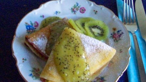 Auf einem dekorativen Teller liegen gefaltete Crepes, überzogen mit einer grünen Kiwi-Soße und Puderzucker, daneben frische Kiwi-Scheiben.