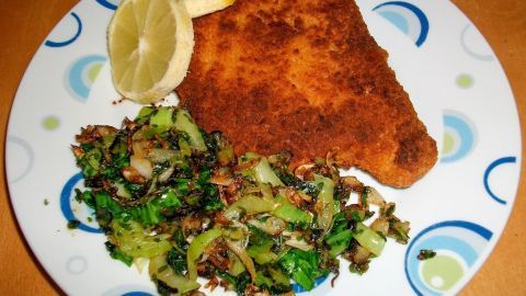 Frittierter Fisch auf einem Teller, umgeben von Pak Choi-Gemüse und Zitronenscheiben, auf einer holzigen Tischfläche.