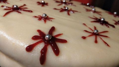 Eine Marzipan-Mohntorte wird dekorativ mit roter Glasur und glänzenden Perlen verziert, auf einem dunklen Untergrund.