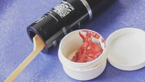 Ein Lippenstift liegt geöffnet auf einer blauen Unterlage, während ein Spatel roten Restlippenstift aus dem Behälter entnimmt.