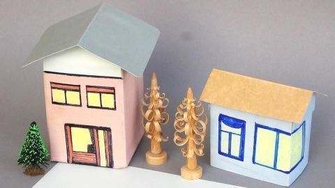 Bunte Tetra Pak-Häuser stehen neben dekorativen Holzbäumen auf einem grauen Hintergrund, perfekt für kreatives Basteln.
