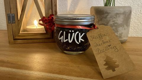 Ein Glas mit "GLÜCK" steht auf einem Tisch, umgeben von einer Laterne und einem Topf mit Pflanze – Kaffee-Zucker-Peeling.