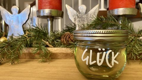 Ein beschriftetes Glas mit dem Wort „GLÜCK“ steht auf einem Holztisch, umgeben von Weihnachtsdekor und Kerzen.