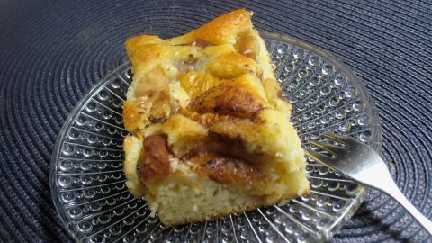 Ein Stück Apfelkuchen auf einem gläsernen Teller, garnierend mit Schmand, steht auf einem dunklen Tischset.
