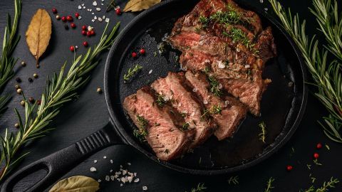 Saftiges, zartes Fleisch liegt in einer gusseisernen Pfanne, umgeben von Kräutern und Gewürzen auf schwarzem Hintergrund.