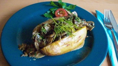 Eine Ofenkartoffel liegt auf einem blauen Teller, gefüllt mit Pilzragout und garniert mit Kräutern und einer Tomatenscheibe.