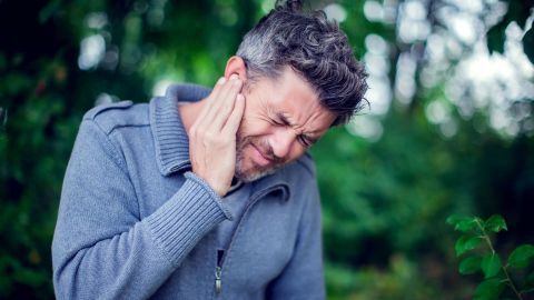 Ein Mann fasst sich an das Ohr, wirkt schmerzhaft und angespannt in einer grünen, natürlichen Umgebung. Tinnitus ist offensichtlich.