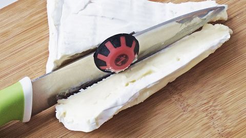 Ein Küchenmesser schneidet durch weichen Käse auf einem Holzbrett, unterstützt von einem Magnetabstreifer zur einfachen Handhabung.